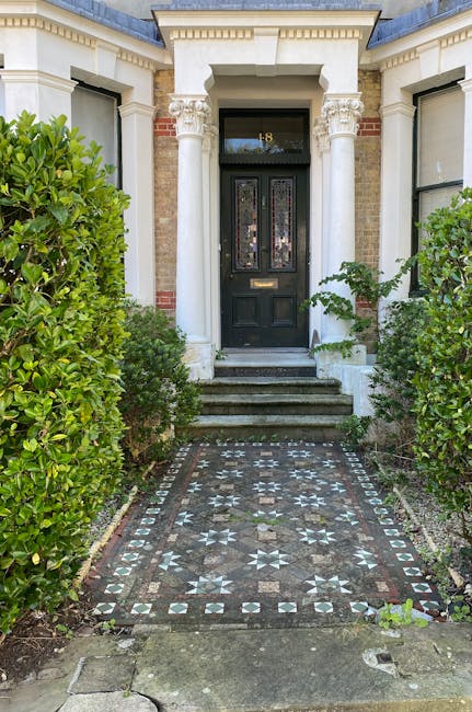 The front garden of a period property features a narrow concrete pathway leading to a white wooden door with a semi-circular arch and a small window above. The garden is bordered by well-maintained, r