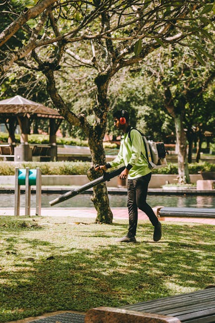 A person wearing a lime green jacket, black pants, and red earmuffs is engaged in garden maintenance, using a leaf blower to clear grass and fallen leaves in a park setting. They are walking across a 