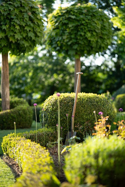 A person wearing a lime green jacket, black pants, and red earmuffs is engaged in garden maintenance, using a leaf blower to clear grass and fallen leaves in a park setting. They are walking across a 